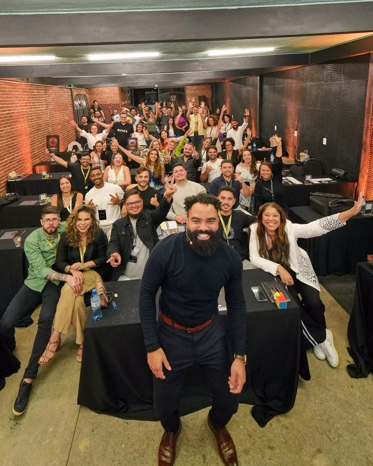 Turma do Ascensão com braços erguidos atrás do mentor sorridente em salão de tijolos