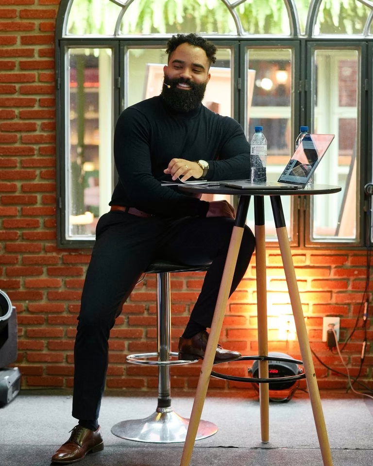 Mentor sentado à mesa alta com laptop, descontraído sob luz quente e parede de tijolos